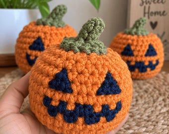 May include: Three small, round, orange crocheted pumpkins with dark blue yarn stitched into triangular eyes and jagged mouths. Each pumpkin features a textured green stem at the top. The pumpkins are crafted with a visible, chunky yarn stitch.