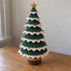 Könnte beinhalten: Ein gehäkelter Weihnachtsbaum mit einem dunkelgrünen, kegelförmigen Körper, weißen, wellenförmigen Rändern, kleinen roten, kugelförmigen Akzenten und einem gelben Stern als Spitze, montiert auf einem braunen, zylindrischen Häkelsockel.