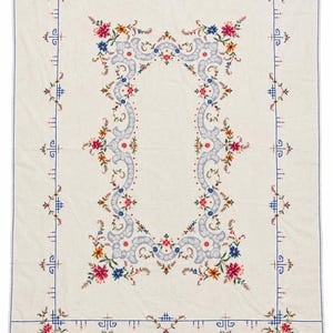 Puede incluir: Un mantel rectangular de color blanco roto que presenta un marco ovalado central de volutas azul claro rodeado de coloridos bordados florales en tonos rosa, azul y amarillo, con un borde azul fino y pequeños detalles geométricos.