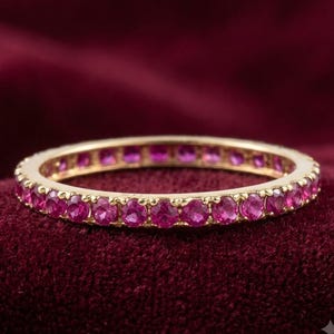 Könnte beinhalten: Ein dünner Goldring mit einer durchgehenden Reihe kleiner, runder, leuchtend pinker Edelsteine, die in einer gemeinsamen Krappenfassung gefasst sind. Der Ring ist kreisförmig und zeigt eine gleichmäßige Anordnung der Steine um das gesamte Band.