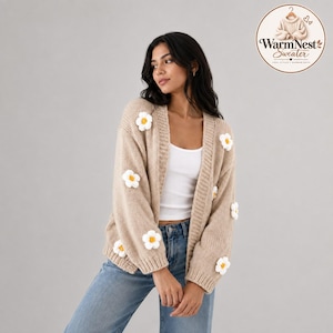 Könnte beinhalten: Eine beigefarbene, vorne offene Strickjacke mit weißen und gelben, dreidimensionalen Blumenapplikationen, die über die Ärmel und Vorderteile verteilt sind. Das Kleidungsstück wird über einem weißen Tanktop und blauen Jeans getragen. Inklusive WarmNest Sweater Logo.