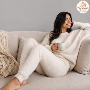 Op de afbeelding: Een crèmekleurige trui met lange mouwen en een bijpassende gebreide broek. De trui heeft een ronde hals en lange mouwen met geribbelde boorden. De broek heeft een aansluitend model met geribbelde enkelboorden. Een keramische mok wordt vastgehouden.