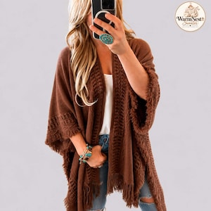 Könnte beinhalten: Eine braune, vorne offene Strickjacke mit strukturiertem Muster an den Vorderkanten und Ärmeln, abgerundet durch Fransen am Saum und an den Ärmelbündchen. Das Kleidungsstück wird über einem weißen Oberteil und einer blauen Jeans getragen.