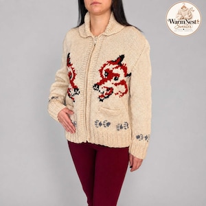 Könnte beinhalten: Eine beige Strickjacke mit Reißverschluss, Kragen, langen Ärmeln und zwei Vordertaschen. Die Jacke zeigt eine rote und schwarze Fuchskopf-Grafik auf der Brust sowie ein kleines blaues Blumenmuster am Saum und an den Ärmelbündchen. Der Text WarmNest Sweater ist sichtbar.