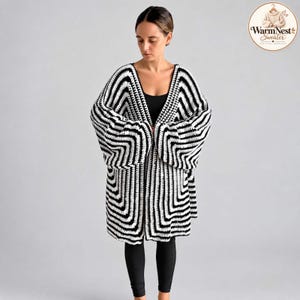 Könnte beinhalten: Eine offene Häkelstrickjacke mit einem markanten schwarz-weißen geometrischen Quadratmuster. Das Kleidungsstück hat weite, lange Ärmel und einen V-Ausschnitt. Ein kreisförmiges Logo in der oberen rechten Ecke zeigt den Text WarmNest Sweater.