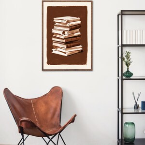 May include: A framed wall art print featuring a stack of books in various shades of brown and cream. The illustration uses visible brushstrokes to depict the layered volumes against a solid dark brown rectangular background with a cream border.