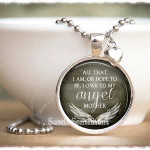 May include: Silver necklace with a round pendant featuring a gray background with white text that reads "All that I am, or hope to be, I owe to my angel mother" with white angel wings. The pendant is attached to a silver chain with a small silver heart charm.