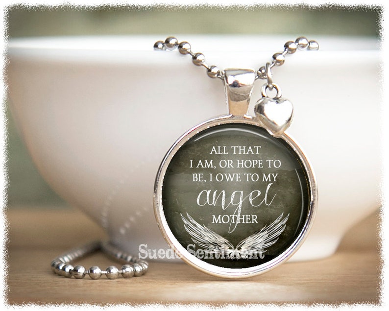 May include: Silver necklace with a round pendant featuring a gray background with white text that reads "All that I am, or hope to be, I owe to my angel mother" with white angel wings. The pendant is attached to a silver chain with a small silver heart charm.