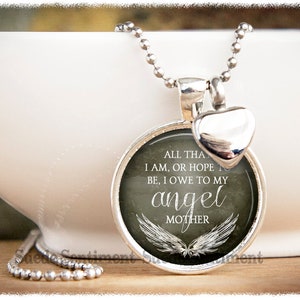 May include: Silver necklace with a round pendant featuring a gray background with white wings and the text "ALL THAT I AM, OR HOPE TO BE, I OWE TO MY ANGEL MOTHER". The pendant is attached to a silver heart charm.