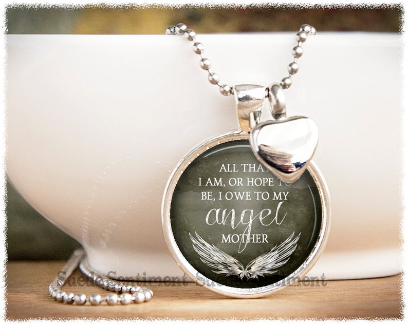 May include: Silver necklace with a round pendant featuring a gray background with white wings and the text "ALL THAT I AM, OR HOPE TO BE, I OWE TO MY ANGEL MOTHER". The pendant is attached to a silver heart charm.