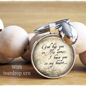 Peut inclure: Un porte-clés en forme d'urne en argent avec un fond blanc et le texte "God has you in His arms: I have you in my heart..."