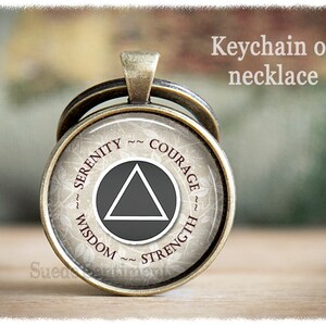 May include: A silver-toned keychain or necklace with a round charm featuring a grey triangle inside a circle. The circle has the words "Serenity", "Courage", "Wisdom", and "Strength" inscribed around it.