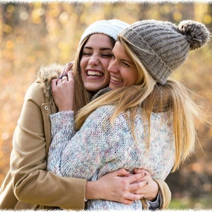 May include: Two women wearing winter hats and sweaters are hugging each other and smiling. They are outdoors in a park with a blurred background of fall foliage.