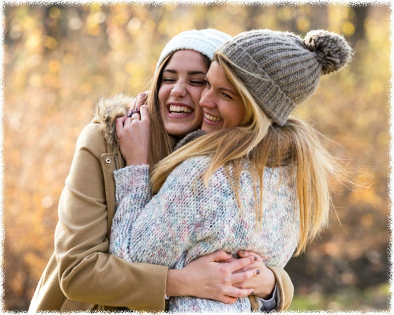 May include: Two women wearing winter hats and sweaters are hugging each other and smiling. They are outdoors in a park with a blurred background of fall foliage.