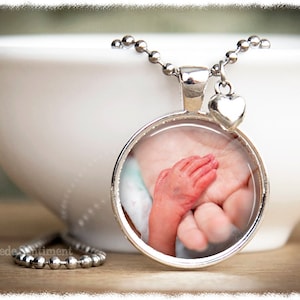 May include: A silver necklace with a round pendant featuring a photo of a baby's hand holding an adult's hand. The pendant has a silver heart charm attached to it.