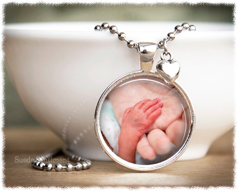 May include: A silver necklace with a round pendant featuring a photo of a baby's hand holding an adult's hand. The pendant has a silver heart charm attached to it.