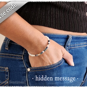 May include: A white, blue and gold beaded bracelet with a hidden message in Morse code. The bracelet is worn on a person's wrist.