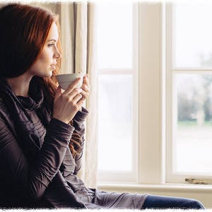 May include: A woman with long red hair sits by a window, holding a white ceramic mug. She is wearing a dark gray long-sleeved shirt and looking out the window.