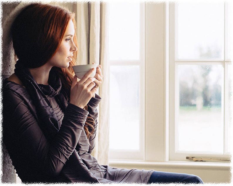 May include: A woman with long red hair sits by a window, holding a white ceramic mug. She is wearing a dark gray long-sleeved shirt and looking out the window.