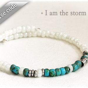 May include: White and turquoise beaded bracelet with silver spacers. The bracelet has the text "I am the storm" written above it.