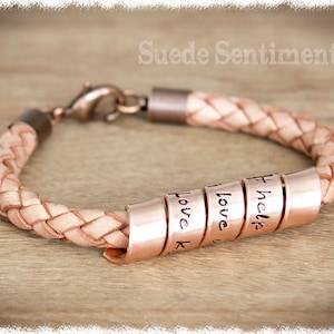 May include: A light brown leather bracelet with a silver clasp and five copper beads. The beads are engraved with the words "I help", "love", "love", "love", and "love K".