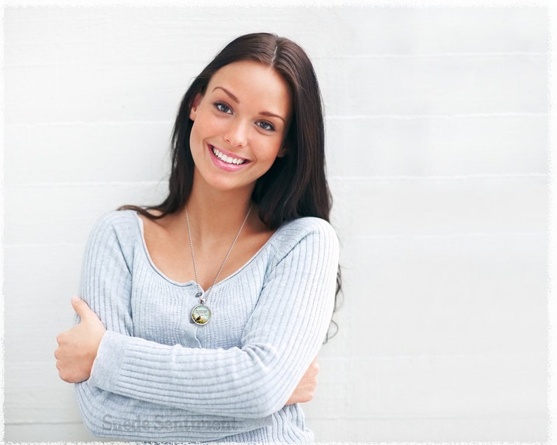 May include: A woman with long brown hair is smiling and wearing a light blue sweater with a silver necklace and a round pendant. The pendant has a nature scene with a tree and a bird.