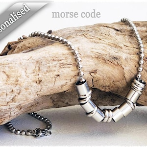 May include: A silver necklace with a chain and a pendant made of multiple silver cylinders. The pendant is in the shape of a U. The necklace is on a piece of driftwood. The text "morse code" is visible above the necklace.