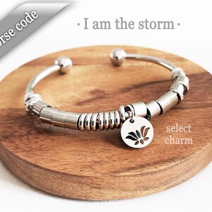 May include: Silver bracelet with a charm featuring a stylised flower design. The bracelet is made of metal beads and has a springy coil section. The text "I am the storm" is visible in the background.