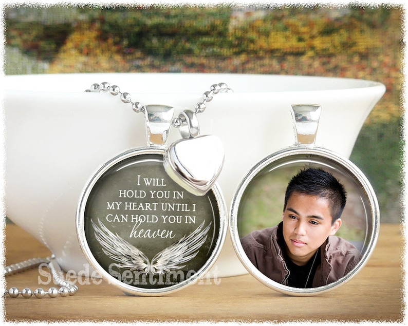May include: A silver necklace with two pendants. One pendant features a photo of a young man, and the other pendant has a white background with the text "I WILL HOLD YOU IN MY HEART UNTIL I CAN HOLD YOU IN heaven" and a pair of white angel wings.