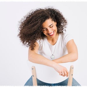 May include: A woman with curly brown hair is sitting on a white chair and smiling. She is wearing a white t-shirt and blue jeans. She is wearing a silver necklace with a round pendant.