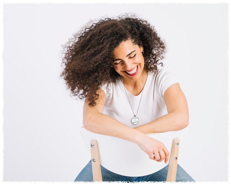 May include: A woman with curly brown hair is sitting on a white chair and smiling. She is wearing a white t-shirt and blue jeans. She is wearing a silver necklace with a round pendant.