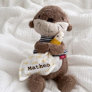 Könnte beinhalten: Ein braunes Plüsch-Otter-Spielzeug in einem dunkelblau-weiß gestreiften Shirt. Der Otter hält einen kleinen weißen Fisch mit gelbem, strukturiertem Körper und einen weißen Stoffbeutel mit gelbem Fischmuster und dem Namen Matheo in brauner Schrift sowie einem roten Etikett.