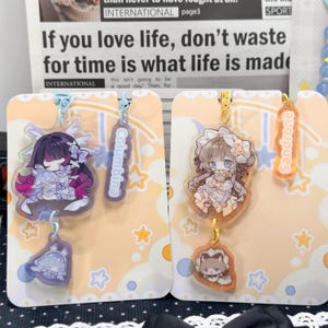 May include: Two acrylic charms featuring anime-style characters on display cards. The left charm depicts a character with dark hair and purple accents labeled Columbina. The right charm shows a character with light hair labeled Sandrone.