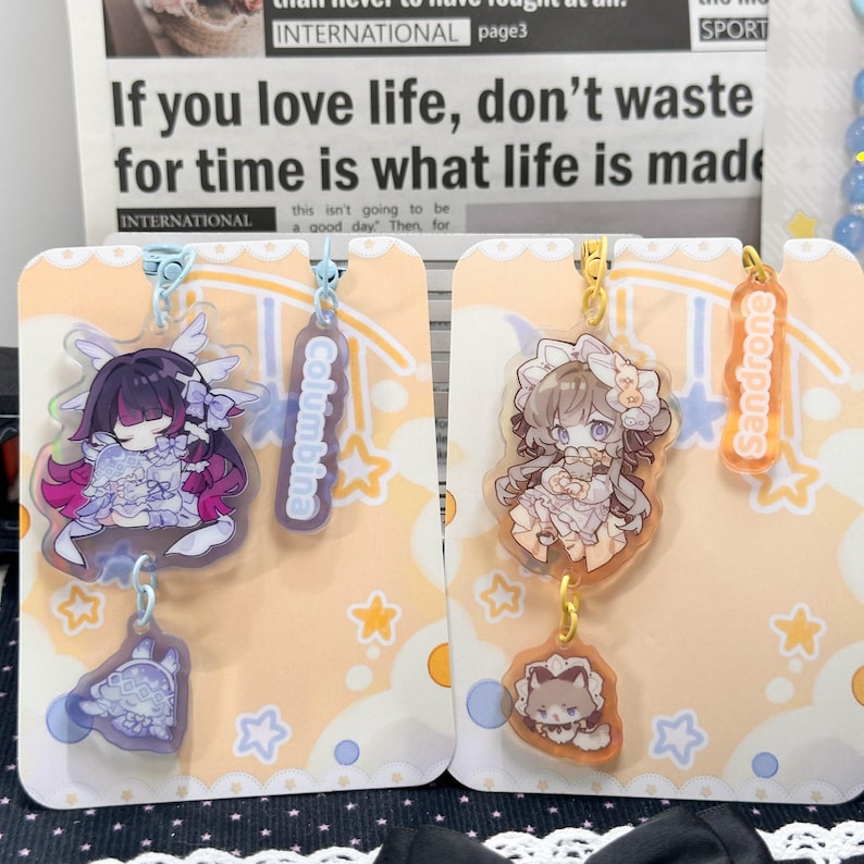 May include: Two acrylic charms featuring anime-style characters on display cards. The left charm depicts a character with dark hair and purple accents labeled Columbina. The right charm shows a character with light hair labeled Sandrone.