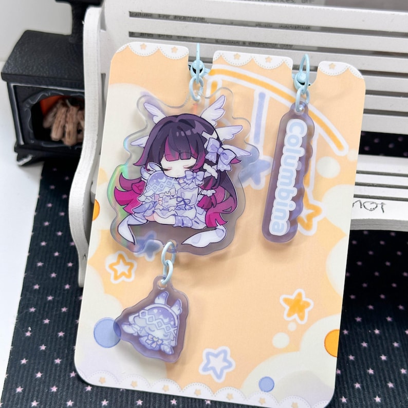 May include: An acrylic charm set featuring a character with dark hair and purple highlights, wearing a white outfit with wings. The set includes a smaller charm of a hooded figure and a rectangular tag with the text Columbina, all on a card.