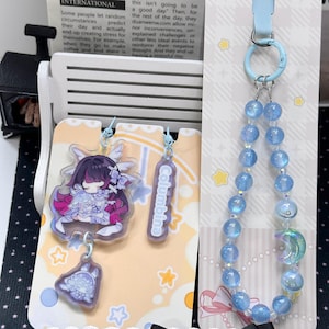 May include: A set of acrylic charms and a beaded phone strap. The charms feature a stylized character with dark hair and a small creature, plus a rectangular tag labeled Columbina. The strap has light blue beads and a crescent moon charm.