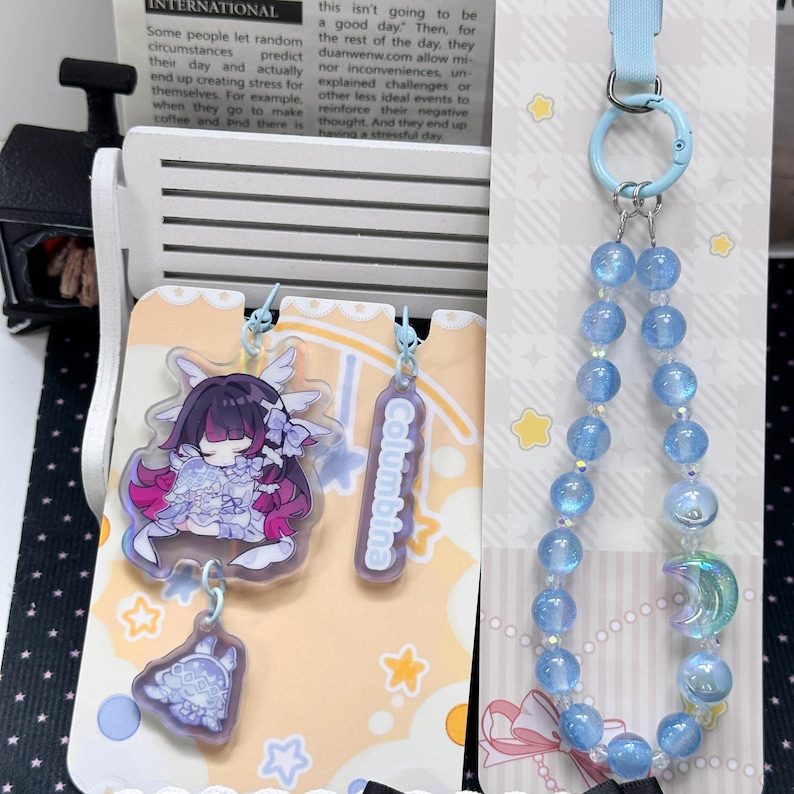 May include: A set of acrylic charms and a beaded phone strap. The charms feature a stylized character with dark hair and a small creature, plus a rectangular tag labeled Columbina. The strap has light blue beads and a crescent moon charm.