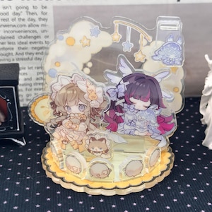 May include: A clear acrylic display stand featuring two illustrated anime-style characters with light brown and dark purple hair, surrounded by small star and cloud motifs, set on a scalloped yellow base with several small circular character icons.