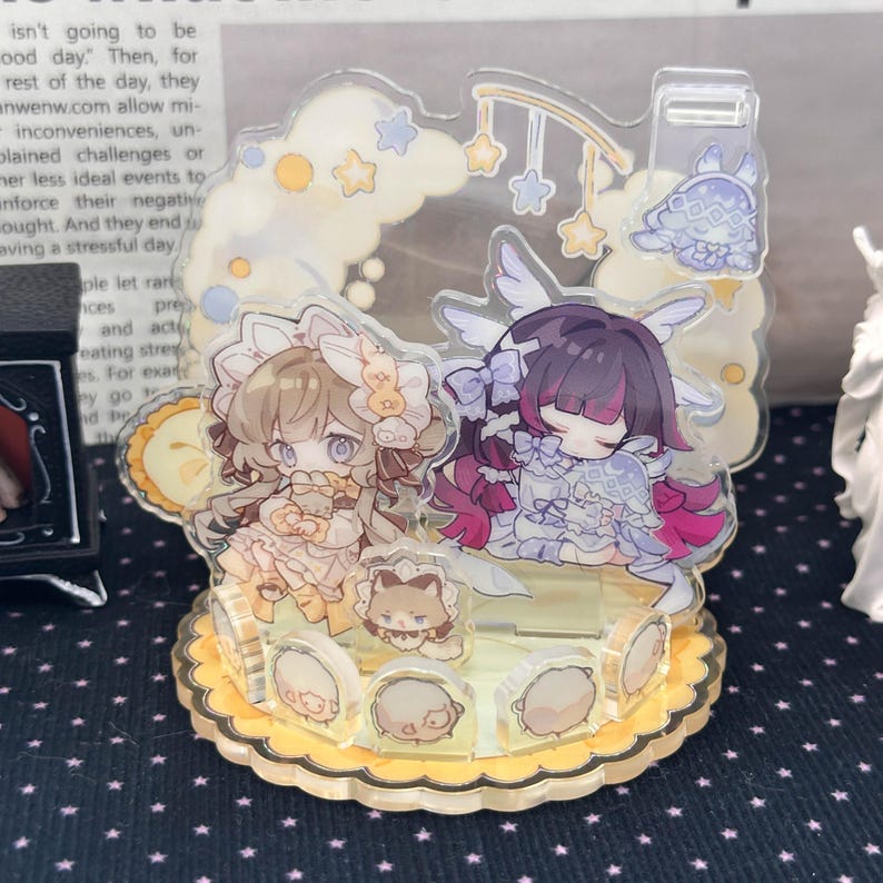 May include: A clear acrylic display stand featuring two illustrated anime-style characters with light brown and dark purple hair, surrounded by small star and cloud motifs, set on a scalloped yellow base with several small circular character icons.