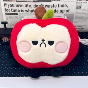 May include: A plush apple toy with a bright red exterior and a cream-colored face. It features embroidered black eyes and a downturned mouth, pink circular cheeks, a brown stem, and a green leaf. The toy has small black arms and legs.