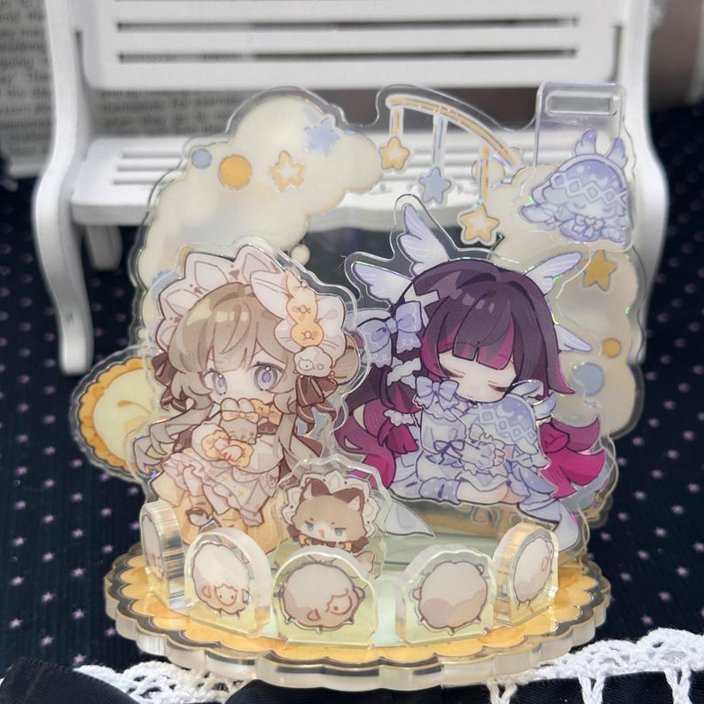 May include: A clear acrylic standee featuring two anime-style characters with pastel hair, one in beige and one in dark purple, surrounded by clouds, stars, and small round creatures on a scalloped base with a light yellow and white color palette.