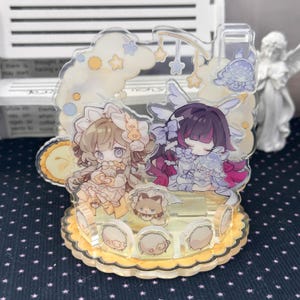 May include: A multi-layered acrylic standee featuring two anime-style characters with light brown and dark purple hair, surrounded by cloud and star motifs. The base is circular with scalloped edges and small circular character icons.