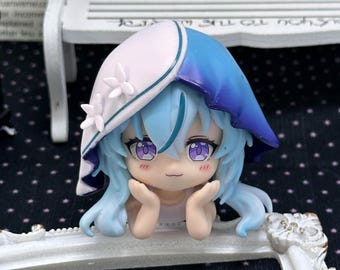 May include: A small anime-style figurine featuring a character with light blue hair, purple eyes, and a small smile. The character wears a white and blue hood adorned with small white flowers and rests their chin on their hands in a resting pose.