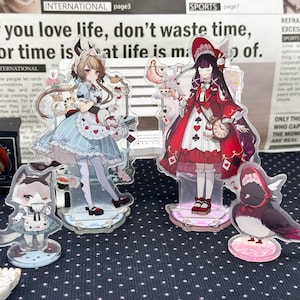 May include: Four acrylic standees featuring anime-style characters and a bird. Two larger figures wear elaborate dresses with card suit motifs, while a smaller cat-like character and a bird in a bonnet stand in front on circular bases.