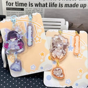 May include: Two acrylic charms featuring anime-style characters on light orange backing cards. The left charm shows a character with dark hair and purple accents labeled Columbina, while the right charm shows a character with light hair labeled Sandrone.