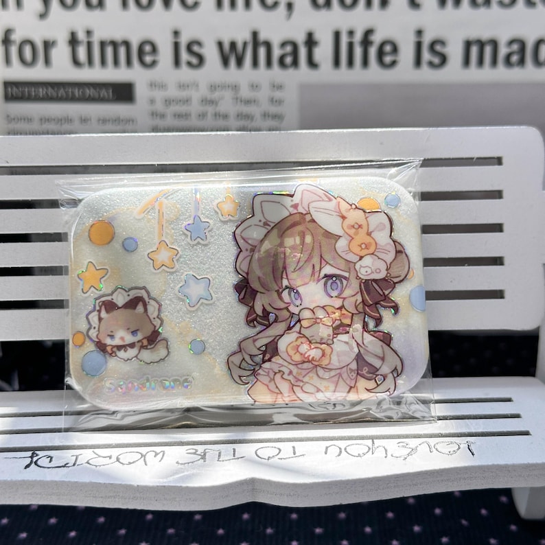 May include: A rectangular acrylic charm featuring a holographic finish with a large anime-style character illustration, a smaller cat-like figure, and various star and circle shapes. The charm is encased in a clear plastic protective sleeve.