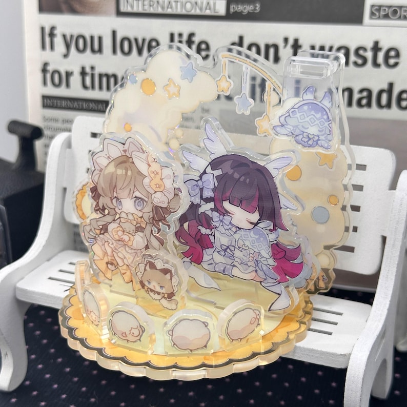 May include: A multi-layered acrylic standee featuring two anime-style characters in frilly outfits, one with light brown hair and one with dark purple hair. The clear acrylic base is decorated with small circular icons and yellow scalloped edges.