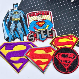 May include: A collection of embroidered fabric patches featuring superhero themes. Included are a full-body Batman figure, a Superman patch with the text THE MAN OF STEEL, two diamond-shaped Superman logos in yellow and purple or red, and a circular patch.