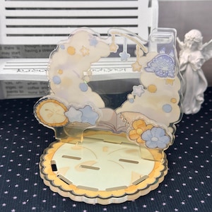 May include: A clear acrylic display stand featuring a printed design of a sleeping character on a cloud, surrounded by stars, flowers, and an open book. The base is circular with a scalloped edge and multiple slots for assembly and item placement.