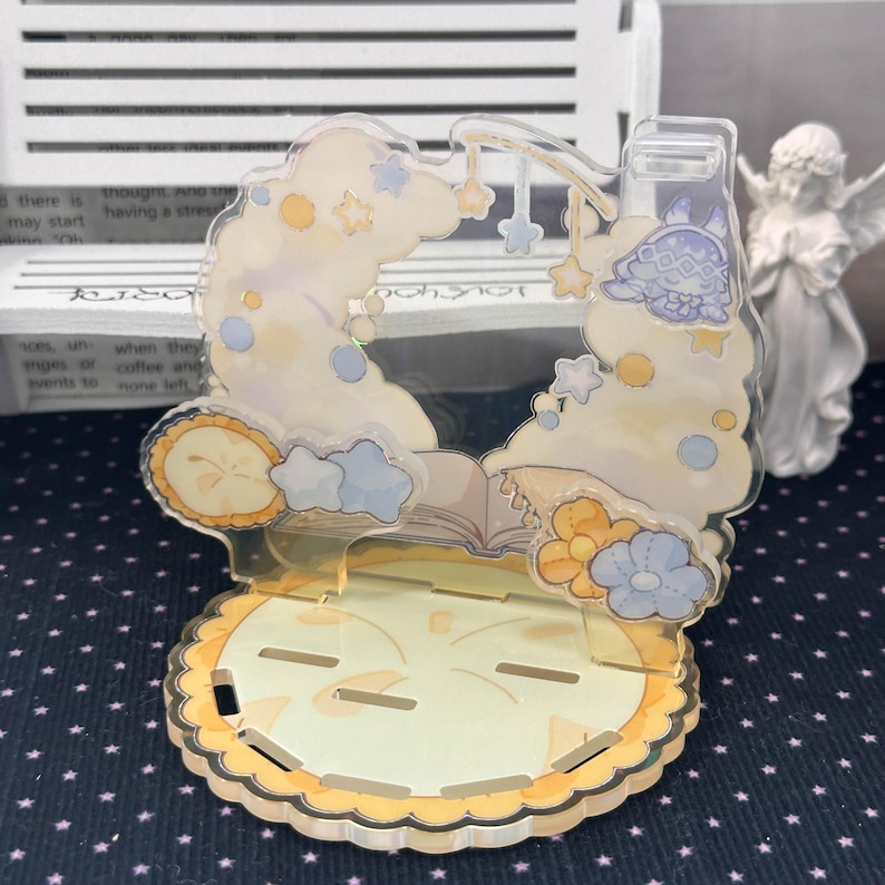 May include: A clear acrylic display stand featuring a printed design of a sleeping character on a cloud, surrounded by stars, flowers, and an open book. The base is circular with a scalloped edge and multiple slots for assembly and item placement.